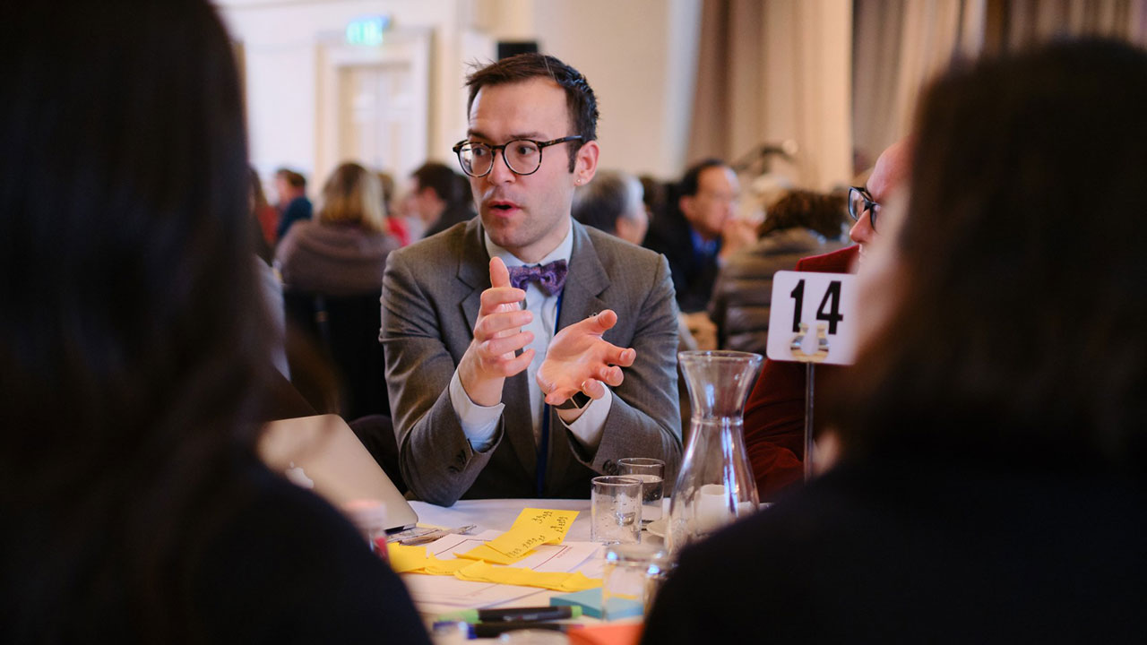 Man wearing glasses and a grey suit, sat across the table and gesturing with his hands.