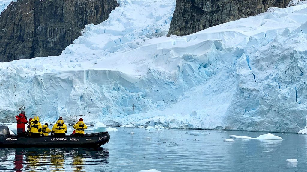 Team of people wearing yellow jackets stand on a small black powerboat, they are looking at white sheets of ice in the ocean.