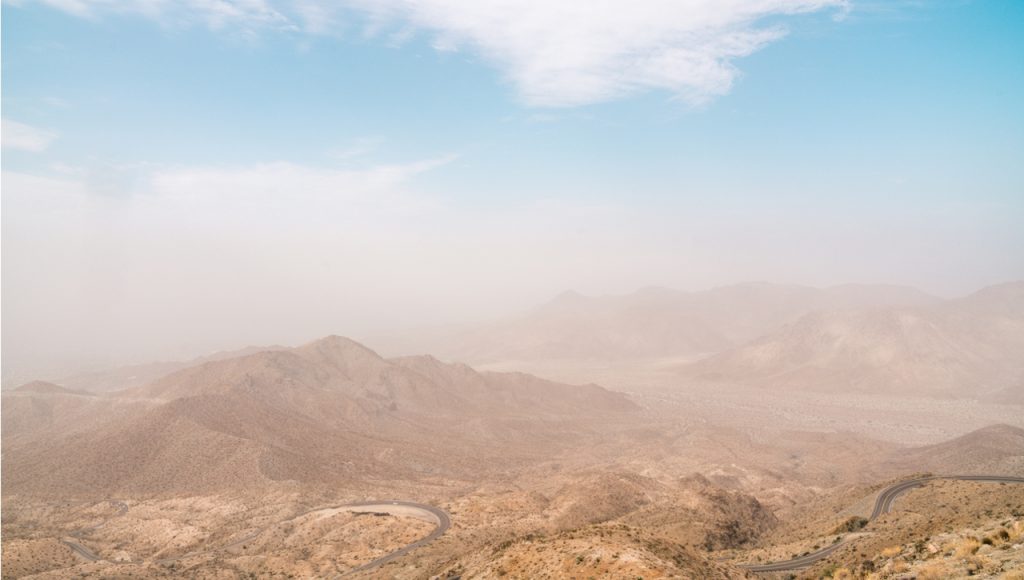 Dust swirls over sand dunes below a light blue sky.