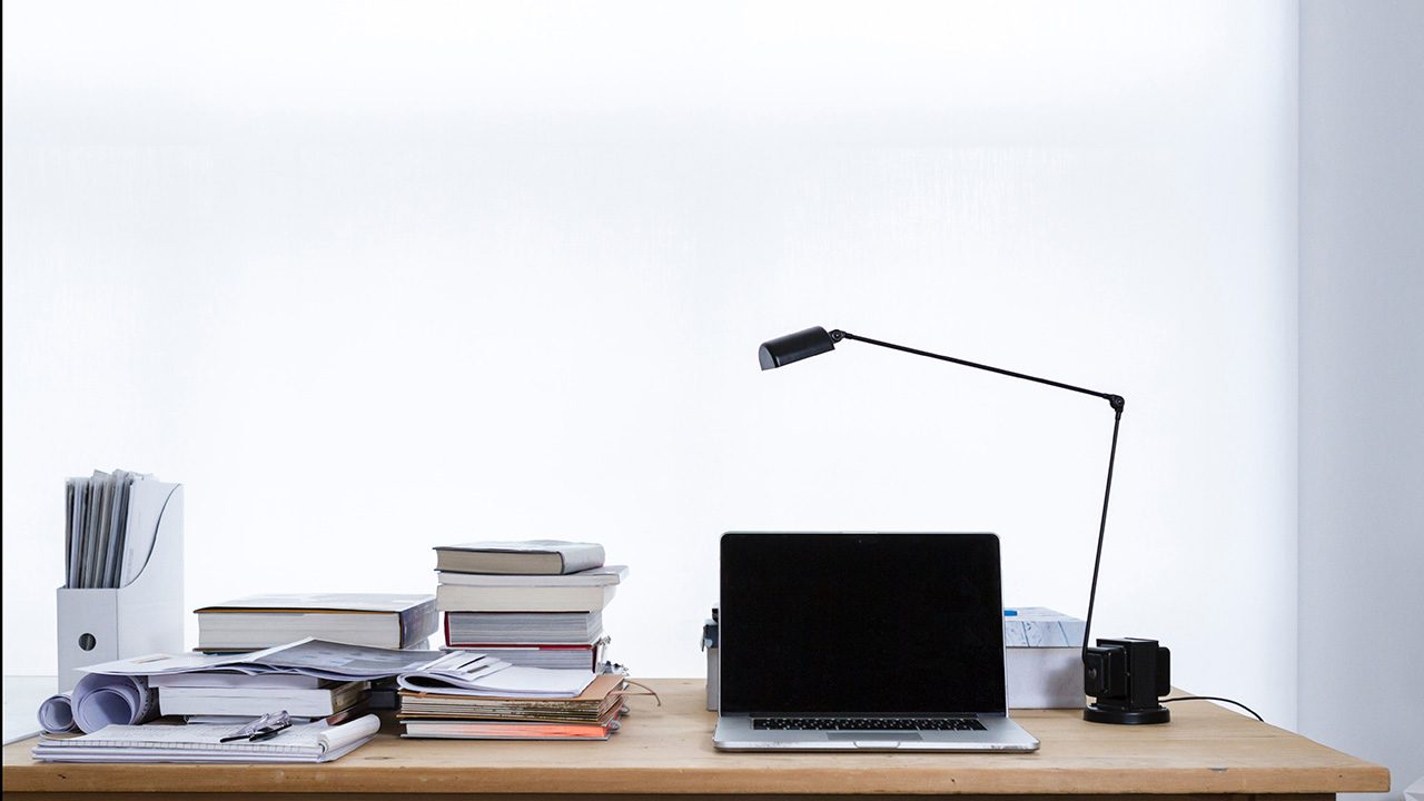 Wooden desk with laptop, reading lamp and piles of books.