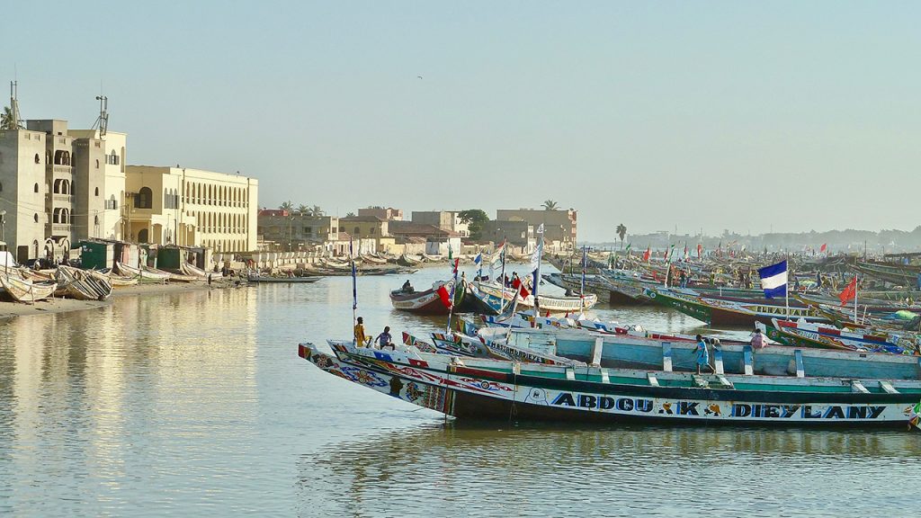 Long fishing boats float in harbour, beside seaside buildings in Senegal