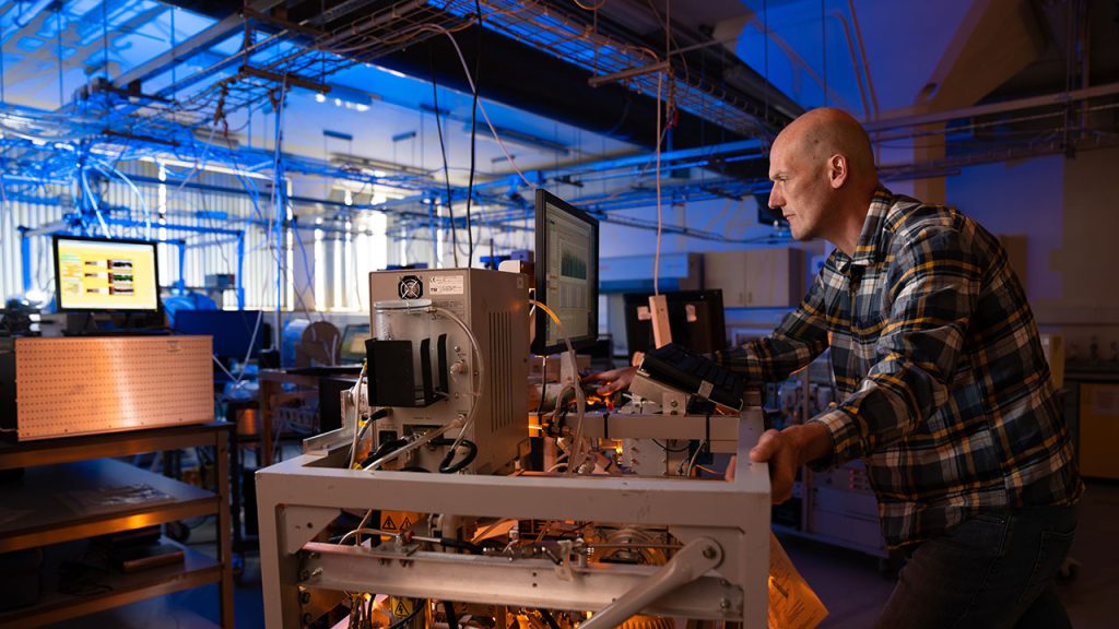 A man in a checked shirt in a laboratory leans over a piece of scientific equipment looking at a computer screen