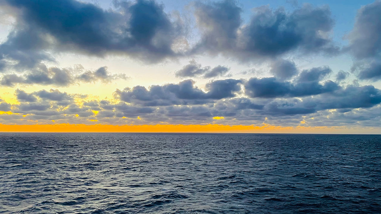 Blue ocean with clouds in the sky and sun setting over the water.
