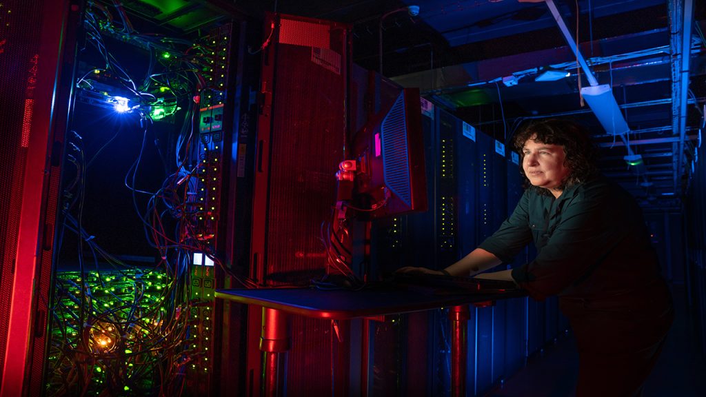 Person with dark curly hair, wearing a green shirt stands at a mobile computer desk in a dark room full of computer equipment.
