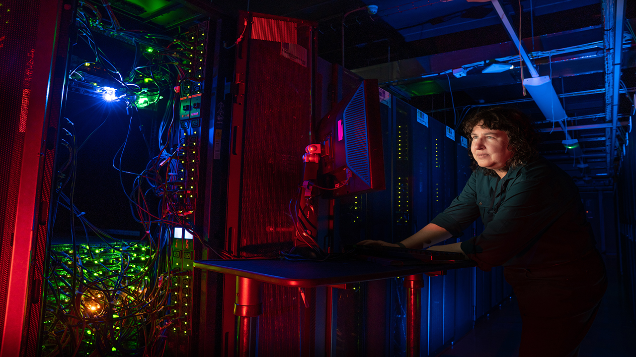 Person with dark curly hair, wearing a green shirt stands at a mobile computer desk in a dark room full of computer equipment.