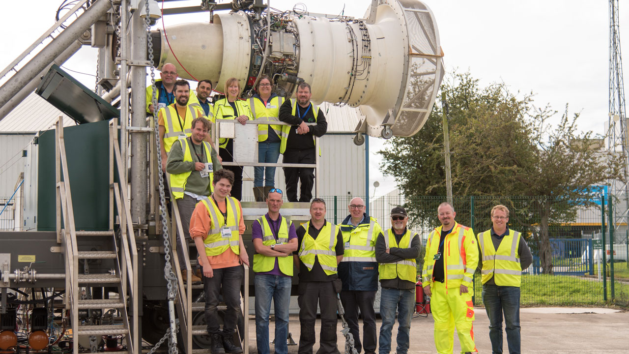 Group of people wearing high visibility jackets stand next to jet engine.