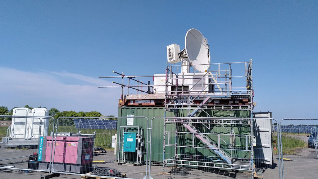 Large white radar on top of green metal container surrounded by metal scaffolding. Blue sky with clouds in background.