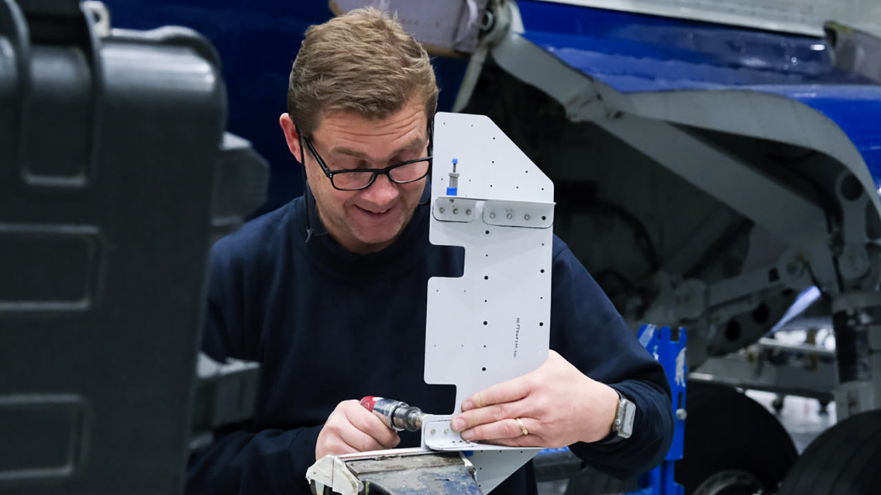 A person uses a tool to modify a metal bracket. They are standing in front of a large research aircraft.