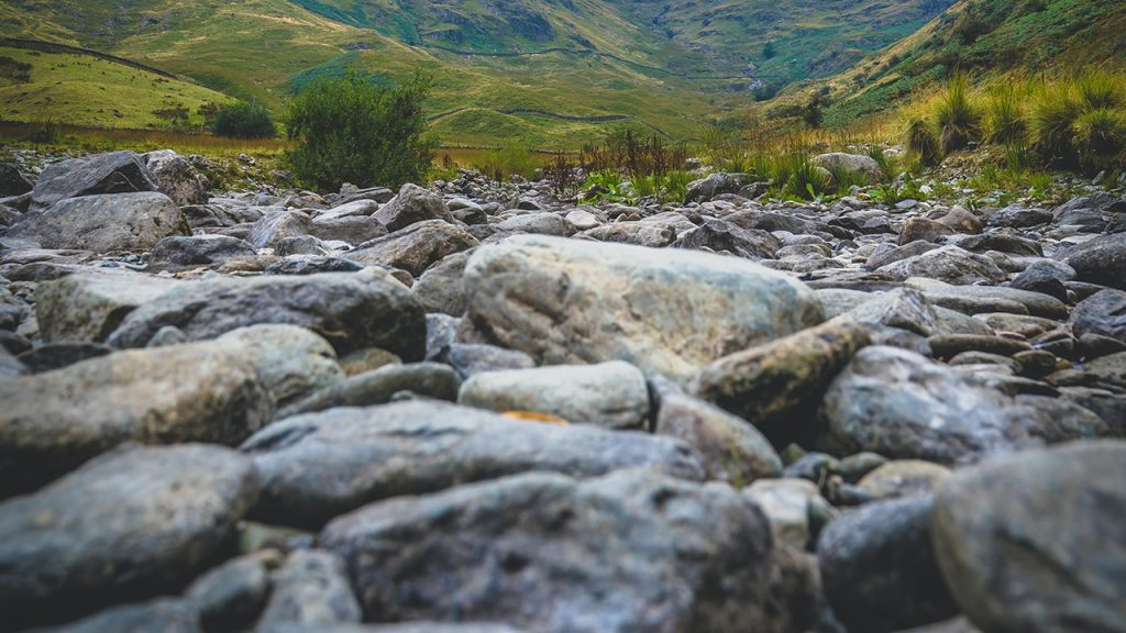 Dry grey pebbles in the foreground, and light green grass and darker green small trees in the background