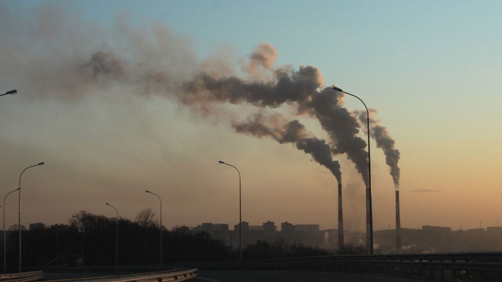 Road and streetlights in foreground. In background, three chimneys are emitting smoke into a dusky sky.