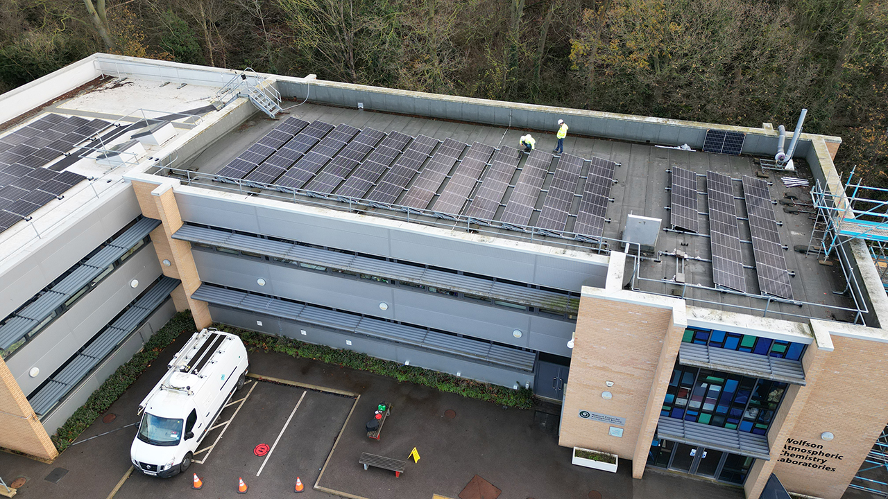Solar panels on the roof of a L shaped building, with two people wearing high visibility jackets inspecting the panels.