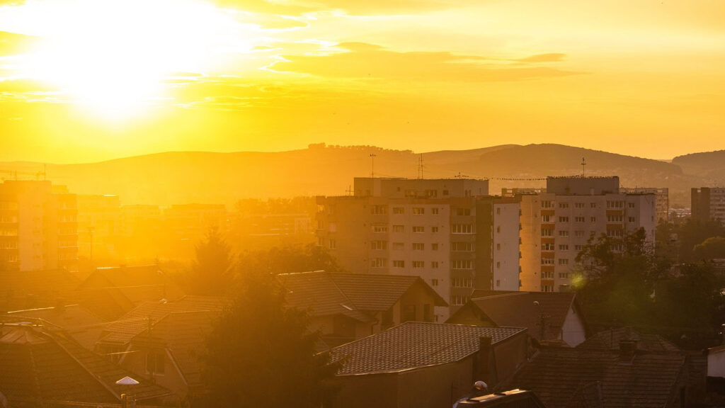 Yellow and orange city skyline with the sun bright in the sky. In the foreground there are houses, trees and flat block towers.