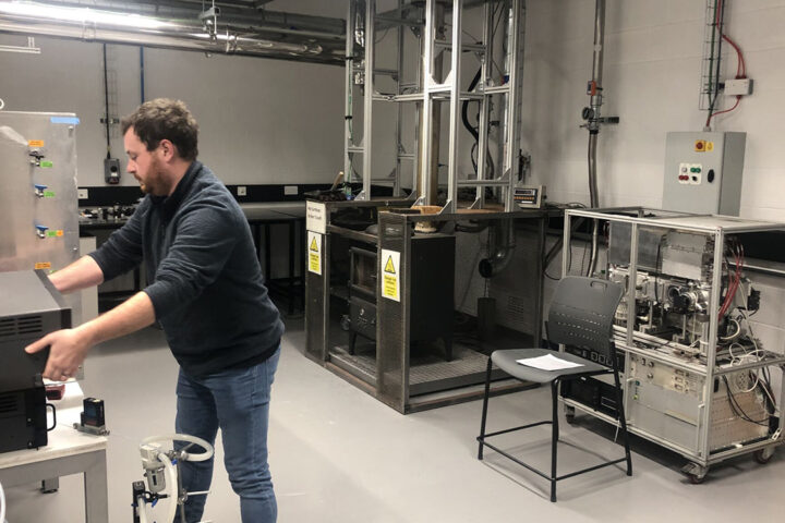 A person with short dark hair, wearing a jumper and jeans, stands facing equipment in a laboratory. The laboratory has a wood burner stove in.