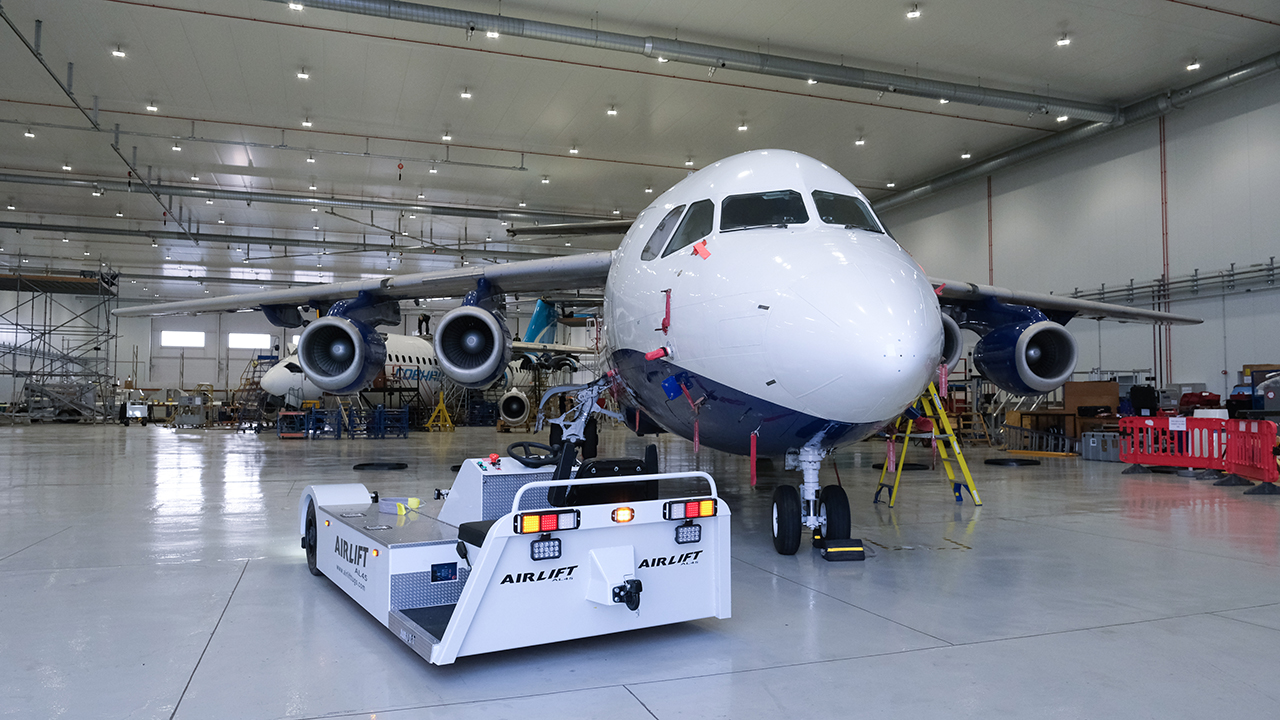An electric tug vehicle sits in front of the nose of a large white and blue aircraft.