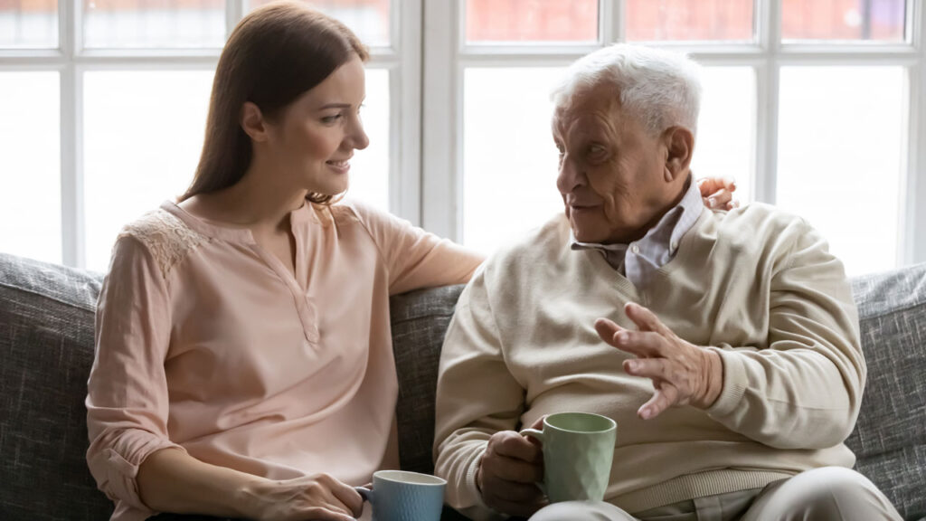 A younger person with long hair and a pink blouse sits on the sofa with an older person with grey hair and a cream jumper. They both hold cups of tea.