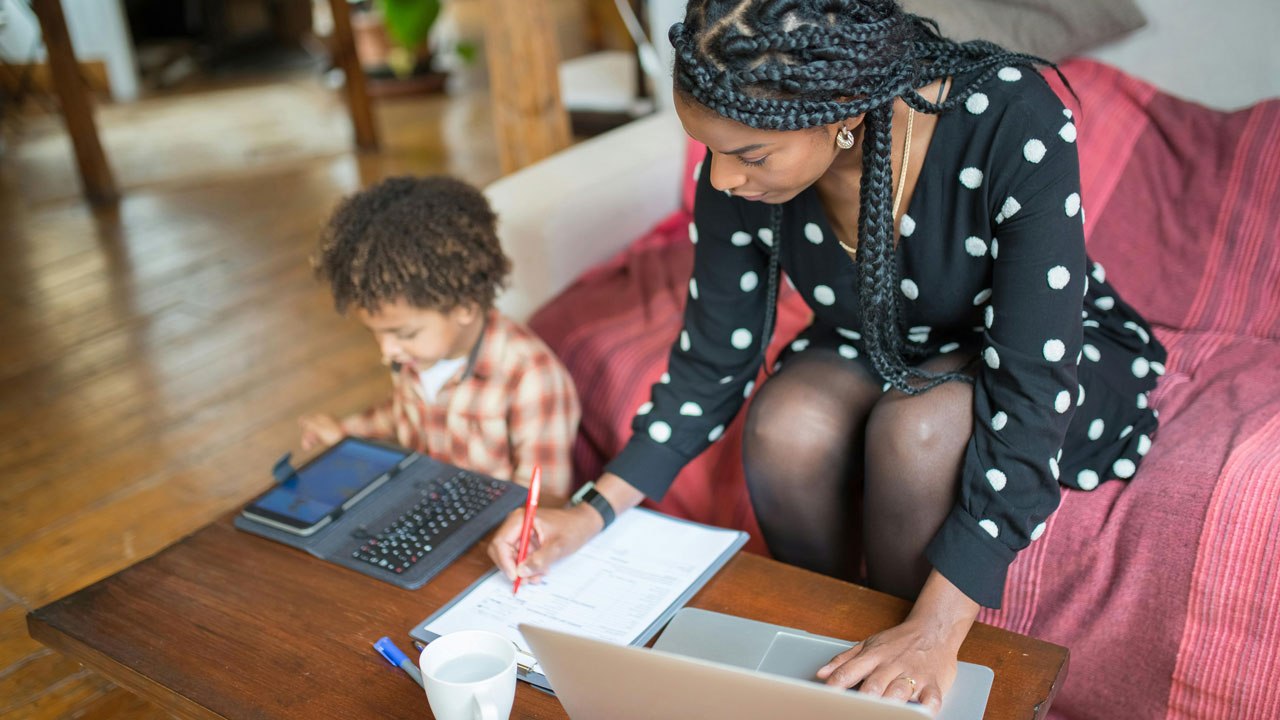 A person sits on the sofa working on their laptop. A child is sitting on the floor next to them playing on a tablet.