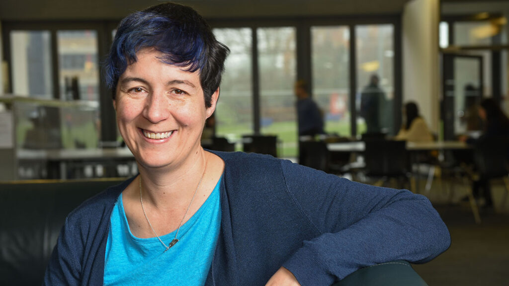 A woman with short dark blue hair, smiling at the camera in a relaxed seated pose, wearing a blue tshirt and cardigan.