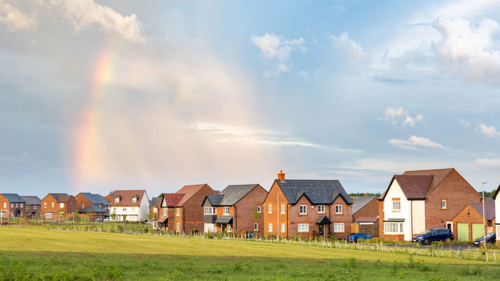 UK housing estate with green grass in foreground and cloudy, blue sky with rainbow in background.