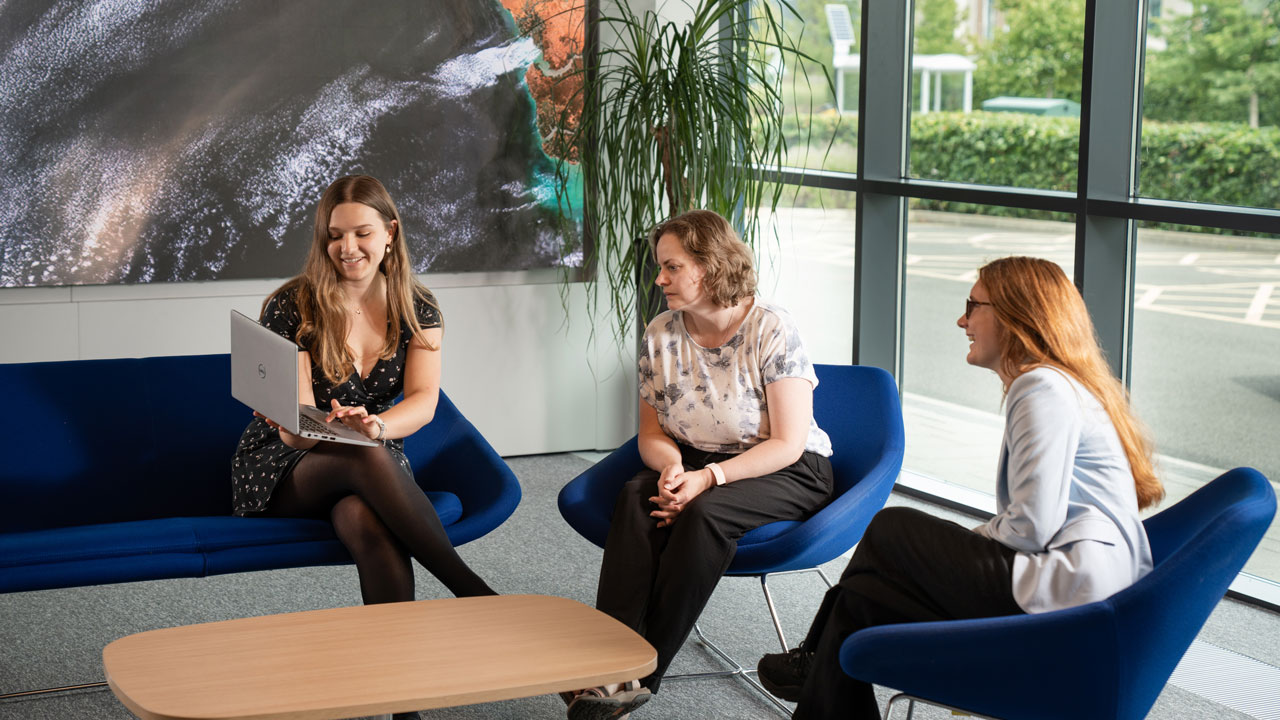 Three people sit in blue chairs. The one on the left is showing something on their laptop.