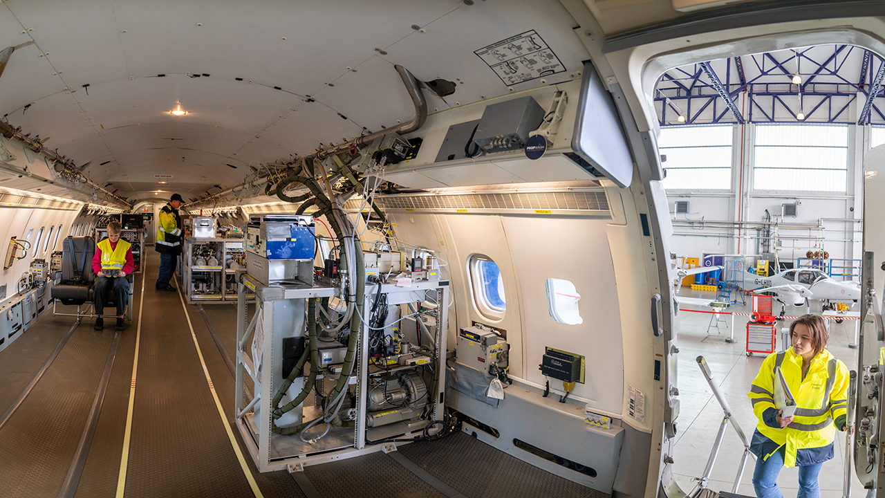 Interior of an aircraft fitted with scientific instruments, with 3 people wearing hi-vis jackets sitting and standing inside.