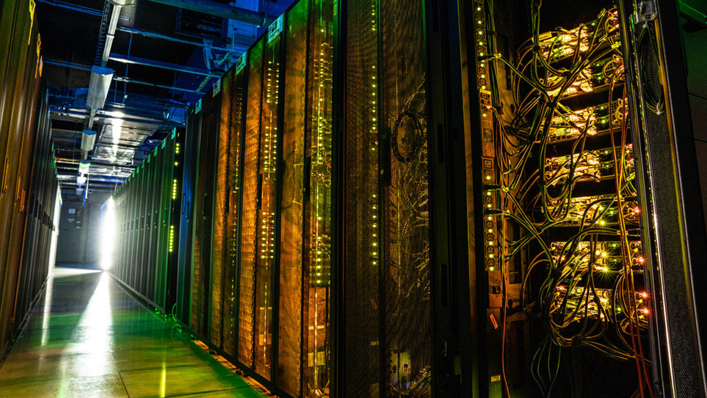 Dark aisle full of racks of computer servers illuminated by small green and blue equipment lights.