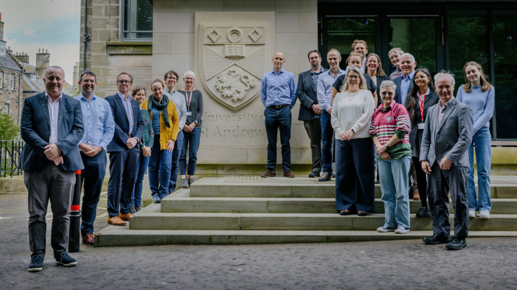 Large group of people stood on stone steps next to a stone University of St Andrews crest on wall.
