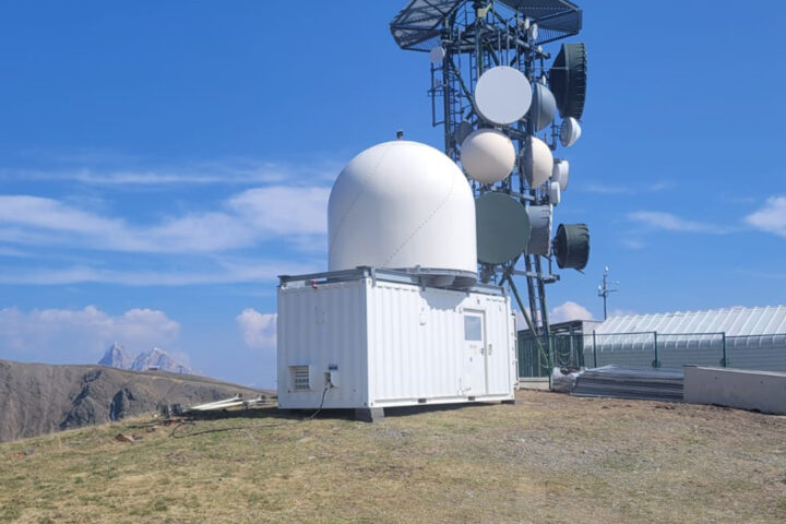 White radar dome on top of a white shipping container, mounted on a grassy hill, with mountains and a blue sky in the background