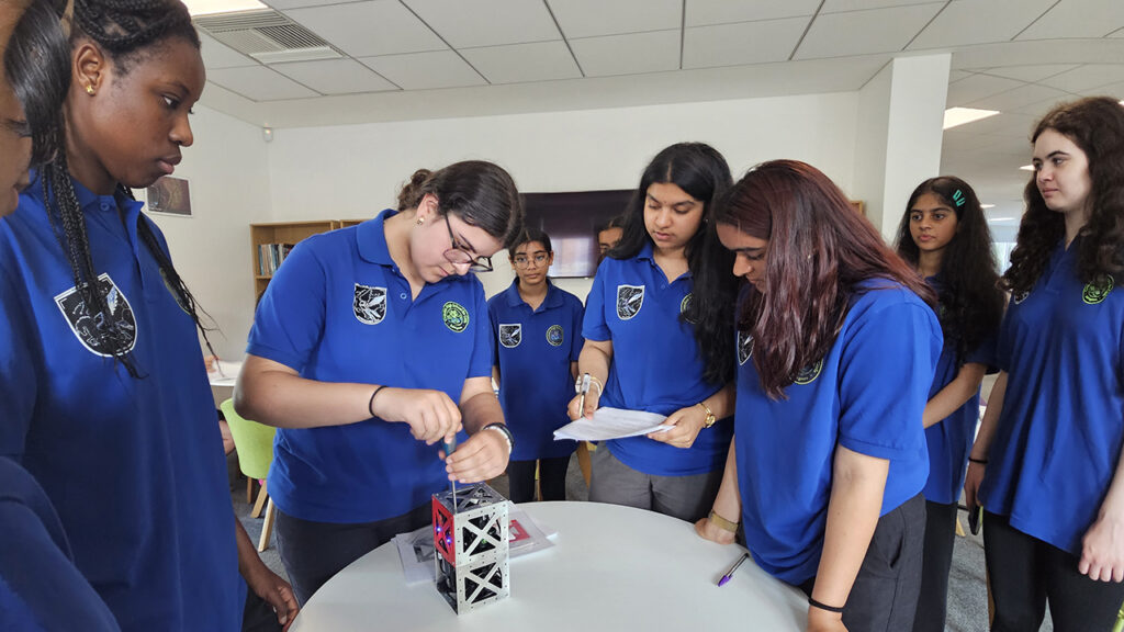 Group of school students wearing blue tshirts, checking a small satellite that's placed on a white table top